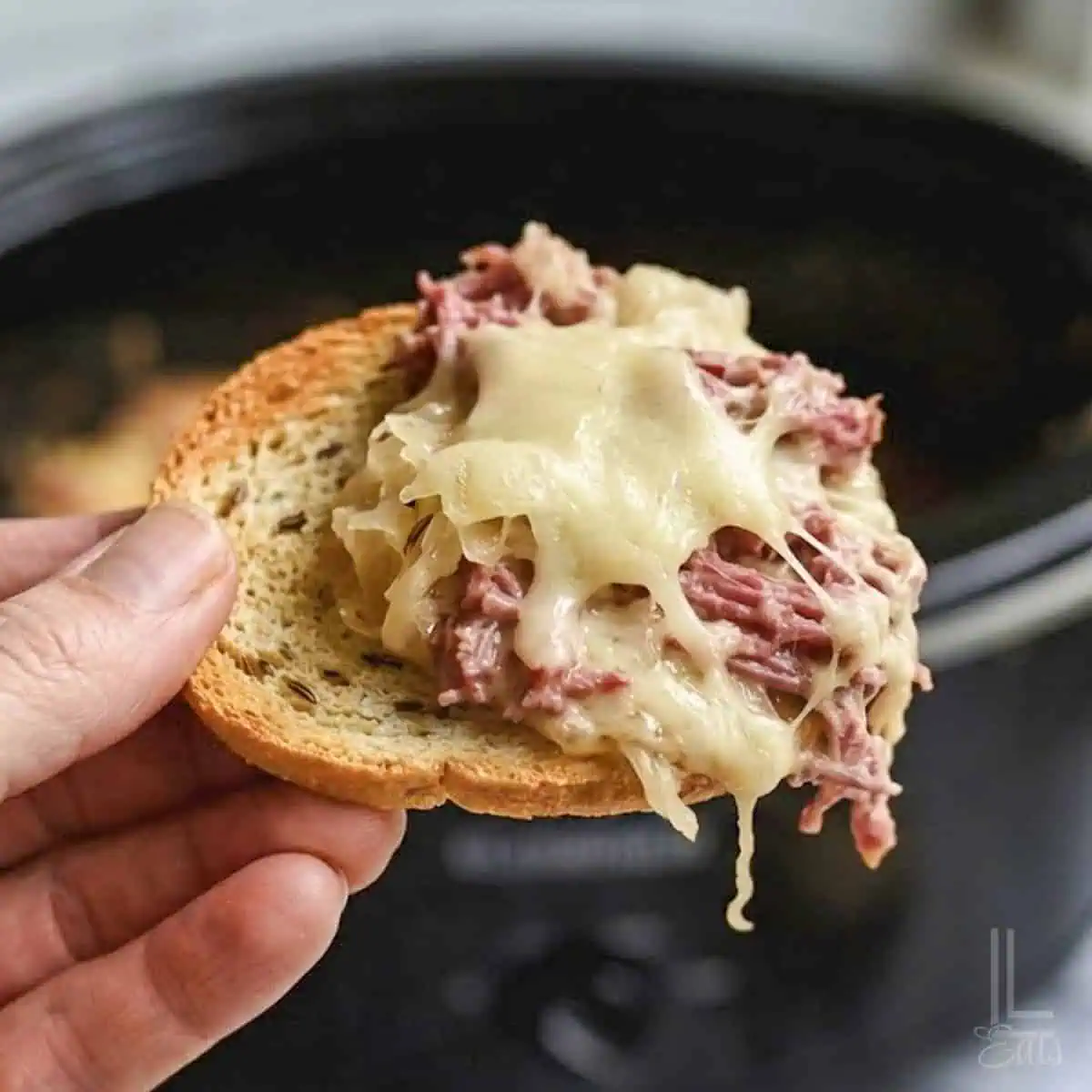 holding a bite of reuben dip on a bagel chip in front of a crock pot.