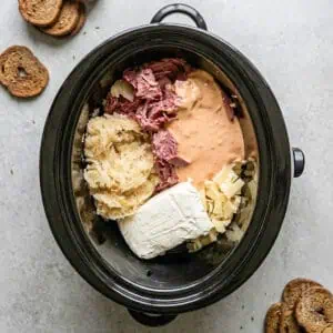making reuben dip in a crock pot; showing corned beef, sauerkraut, thousand island dressing, cream cheese, and swiss cheese.