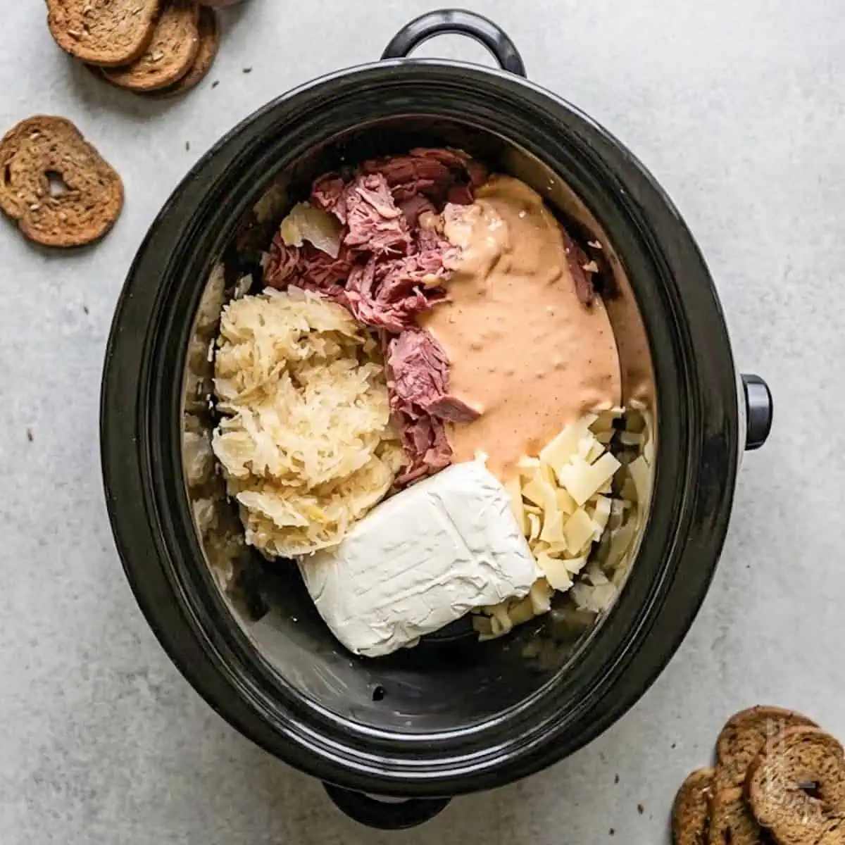 making reuben dip in a crock pot; showing corned beef, sauerkraut, thousand island dressing, cream cheese, and swiss cheese.