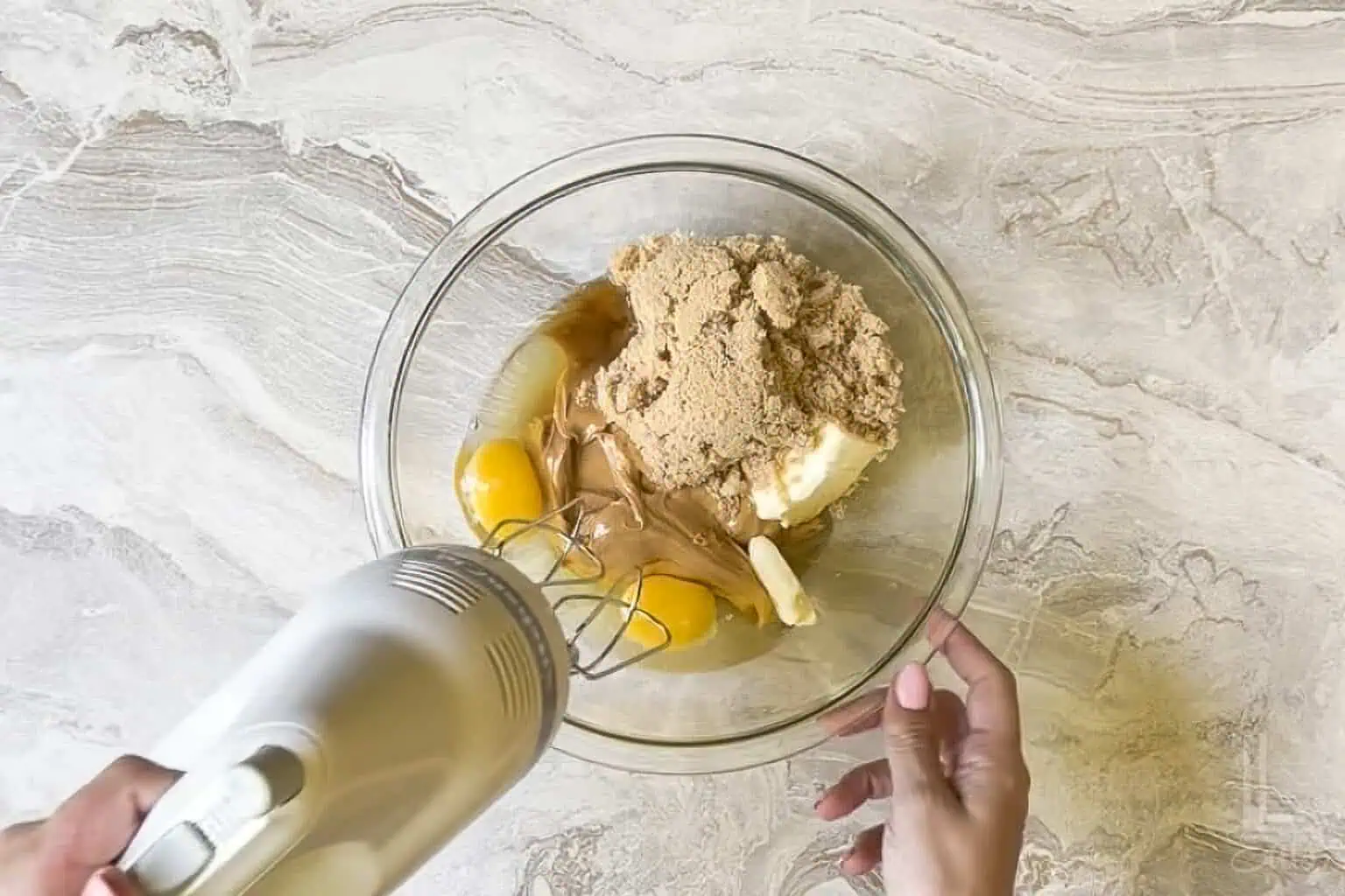 mixing wet ingredients (brown sugar, eggs, vanilla extract, butter) in a large glass bowl with a hand mixer.