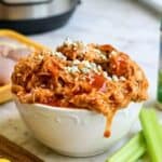 Close-up of shredded buffalo chicken in a white bowl with celery sticks and butter in the background.