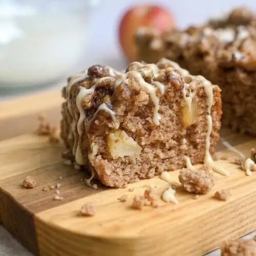 a piece of apple spice cake on a cutting board, surrounded by crumbs, drizzled with glaze and caramel sauce.