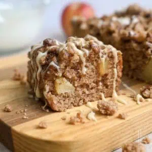 a piece of apple spice cake on a cutting board, surrounded by crumbs, drizzled with glaze and caramel sauce.