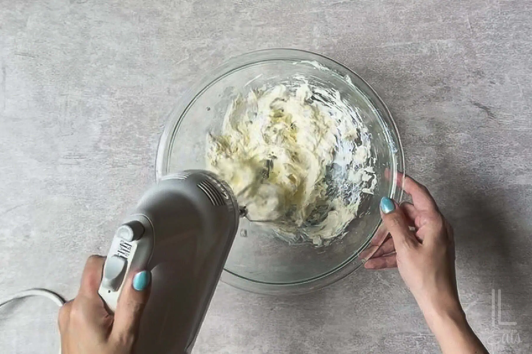 using a hand mixer to whip cream cheese in a glass bowl.