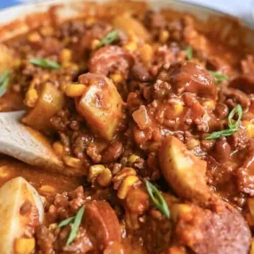 a close up of cowboy stew with a wooden spoon in the pot, showing potatoes, corn, and sausage.