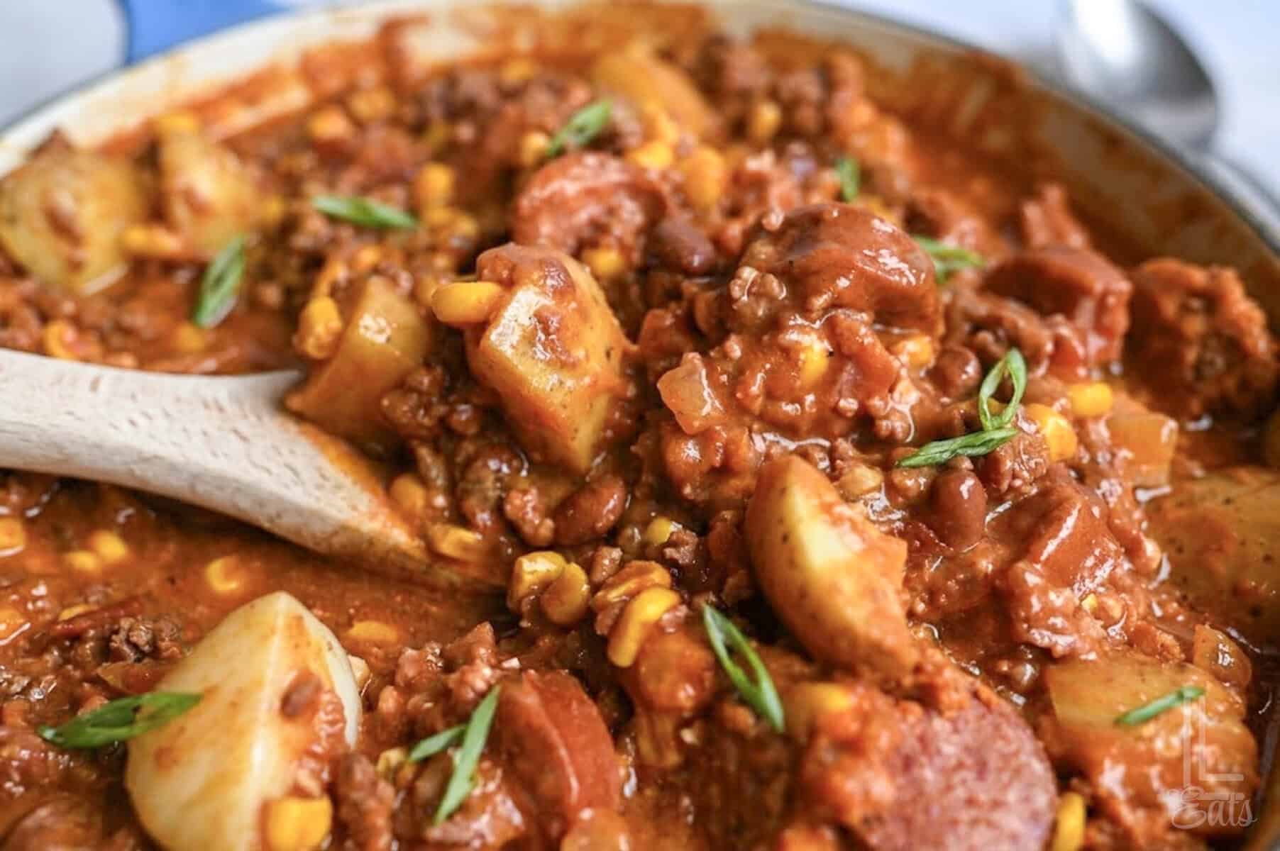 a close up of cowboy stew with a wooden spoon in the pot, showing potatoes, corn, and sausage.