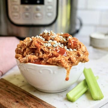bowl of pulled buffalo chicken in front of an instant pot, covered in hot sauce and blue cheese, next to celery sticks.