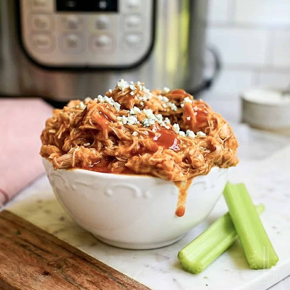 bowl of pulled buffalo chicken in front of an instant pot, covered in hot sauce and blue cheese, next to celery sticks.
