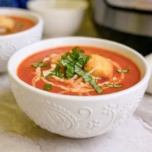 bowls of tomato soup in front of an instant pot, garnished with croutons, parmesan cheese, and cream.