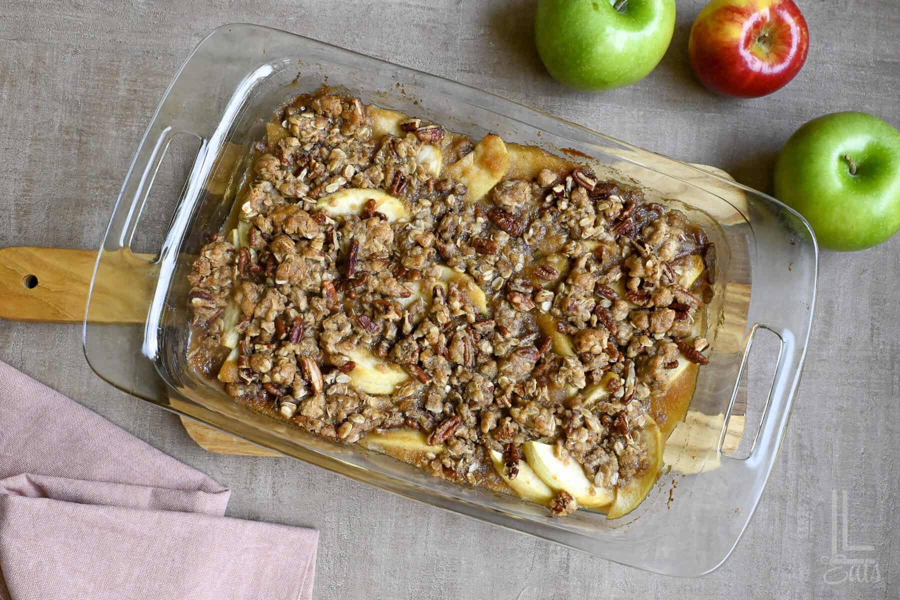 Apple crisp with an oat pecan topping in glass baking dish on table with apples.