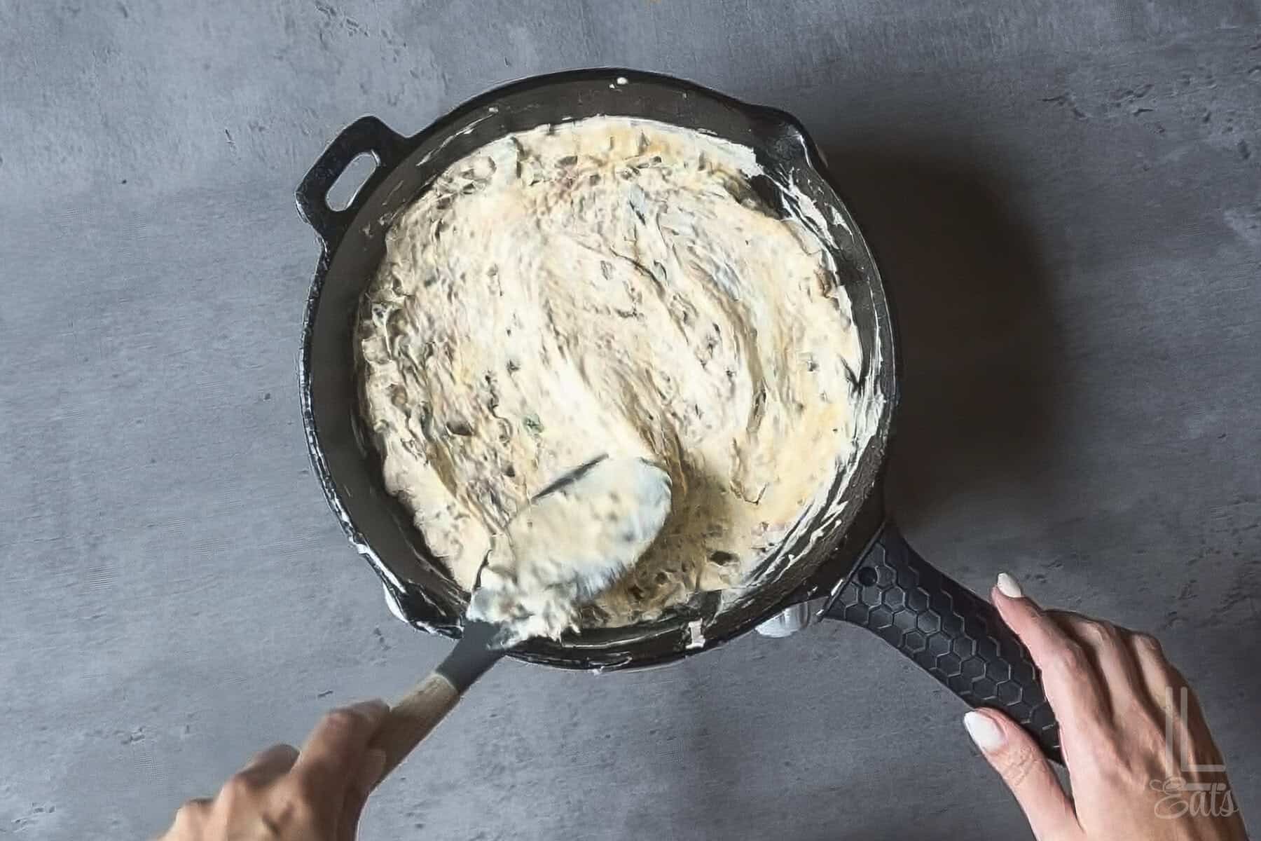 Smoothing creamy jalapeño popper dip into an even layer in a cast-iron skillet.