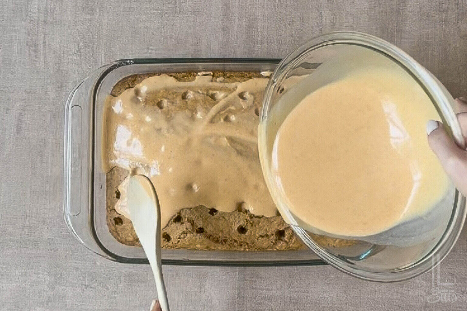 Pouring creamy cheesecake pumpkin pudding over pumpkin poke cake in a glass dish.