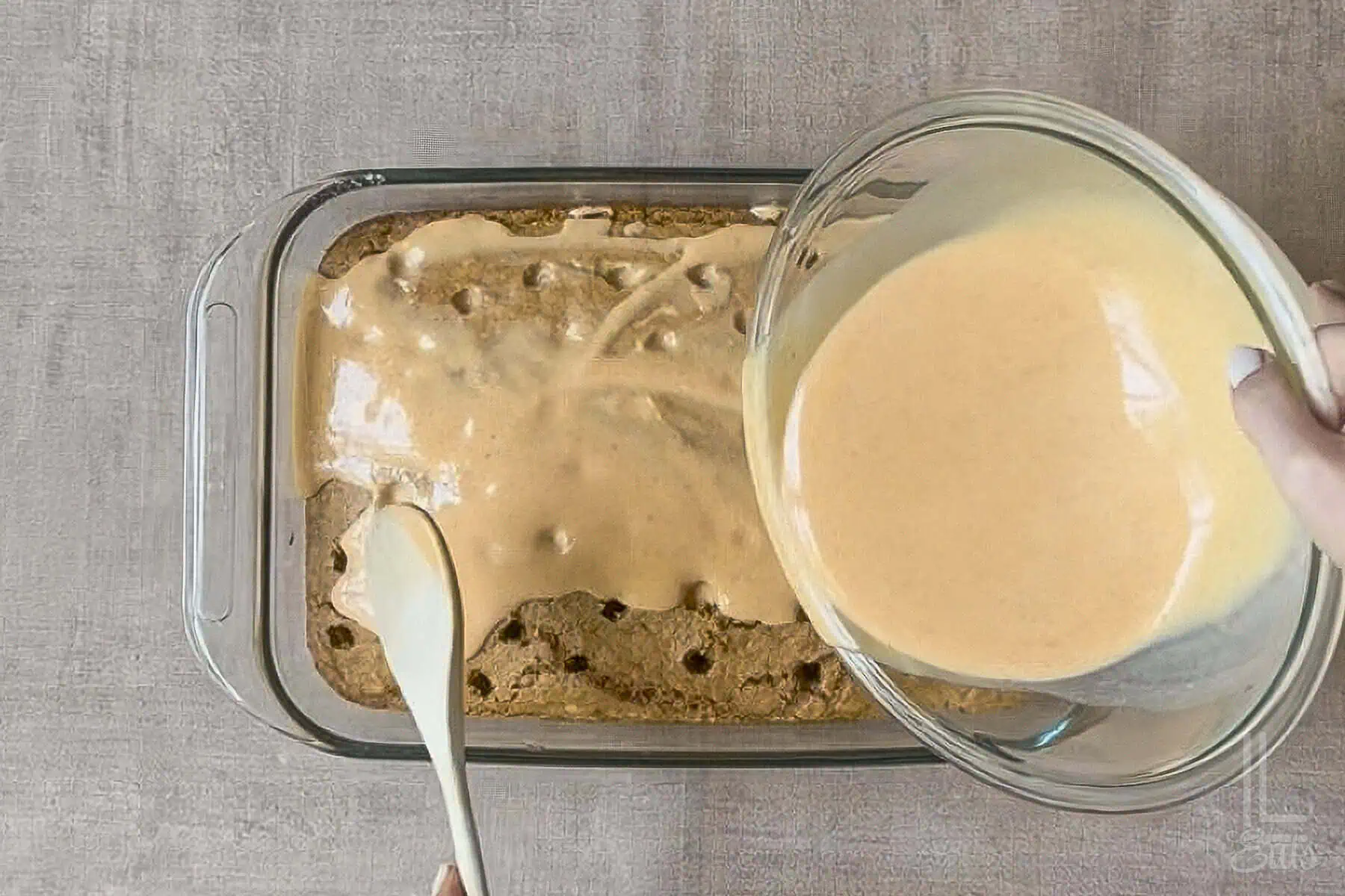 Pouring creamy cheesecake pumpkin pudding over pumpkin poke cake in a glass dish.