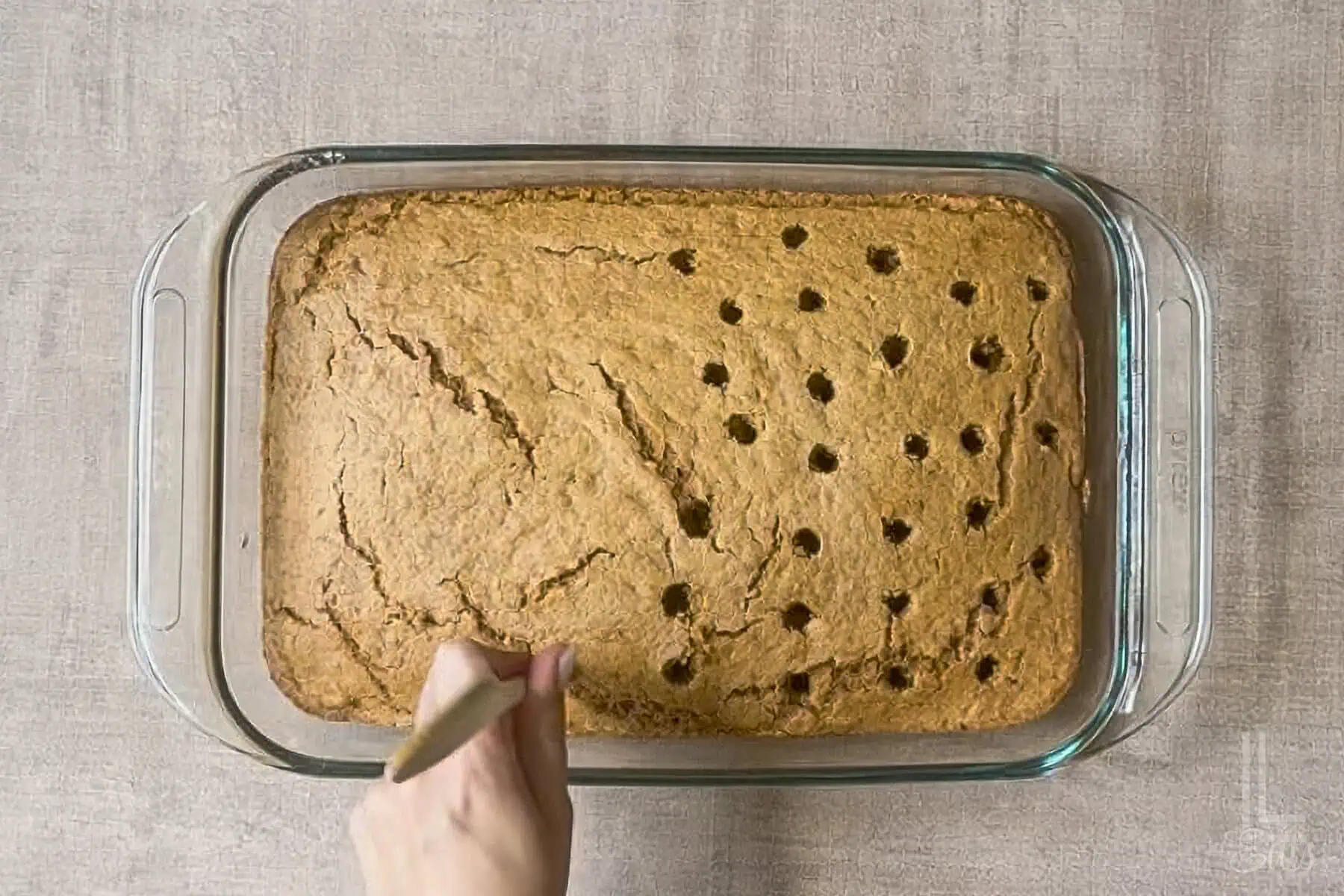 Baked pumpkin poke cake in glass dish, poking holes with a wooden spoon handle.
