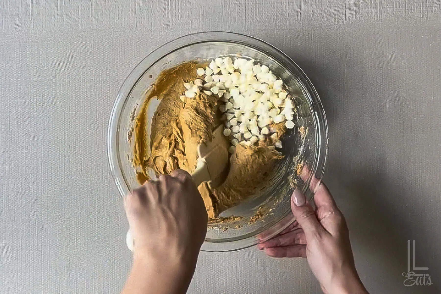 Mixing white chocolate chips into pumpkin cookie dough in a glass bowl for baking.