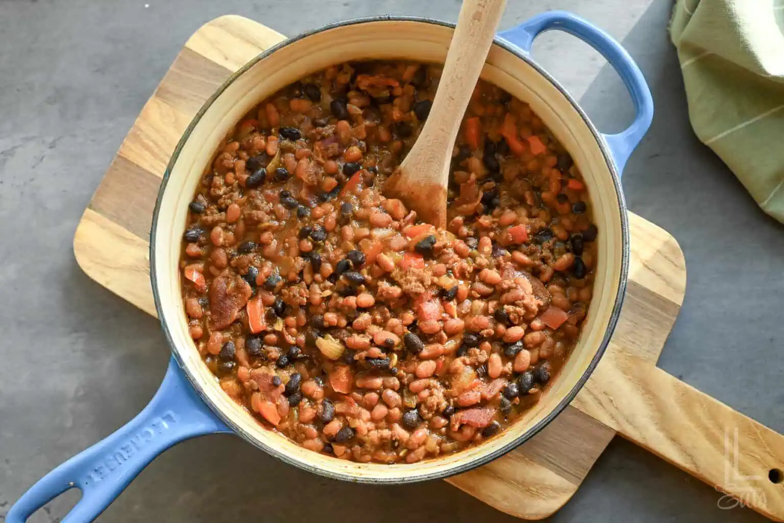 Hearty cowboy baked beans with tomatoes and spices in a blue pot on a wooden board.