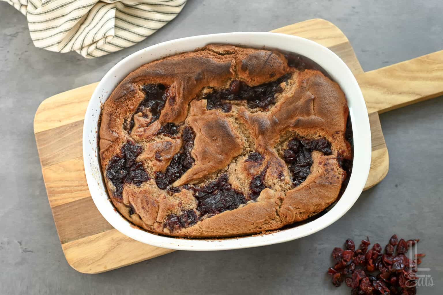 Fresh cranberry cobbler in a white baking dish on a wooden board with dried cranberries nearby.