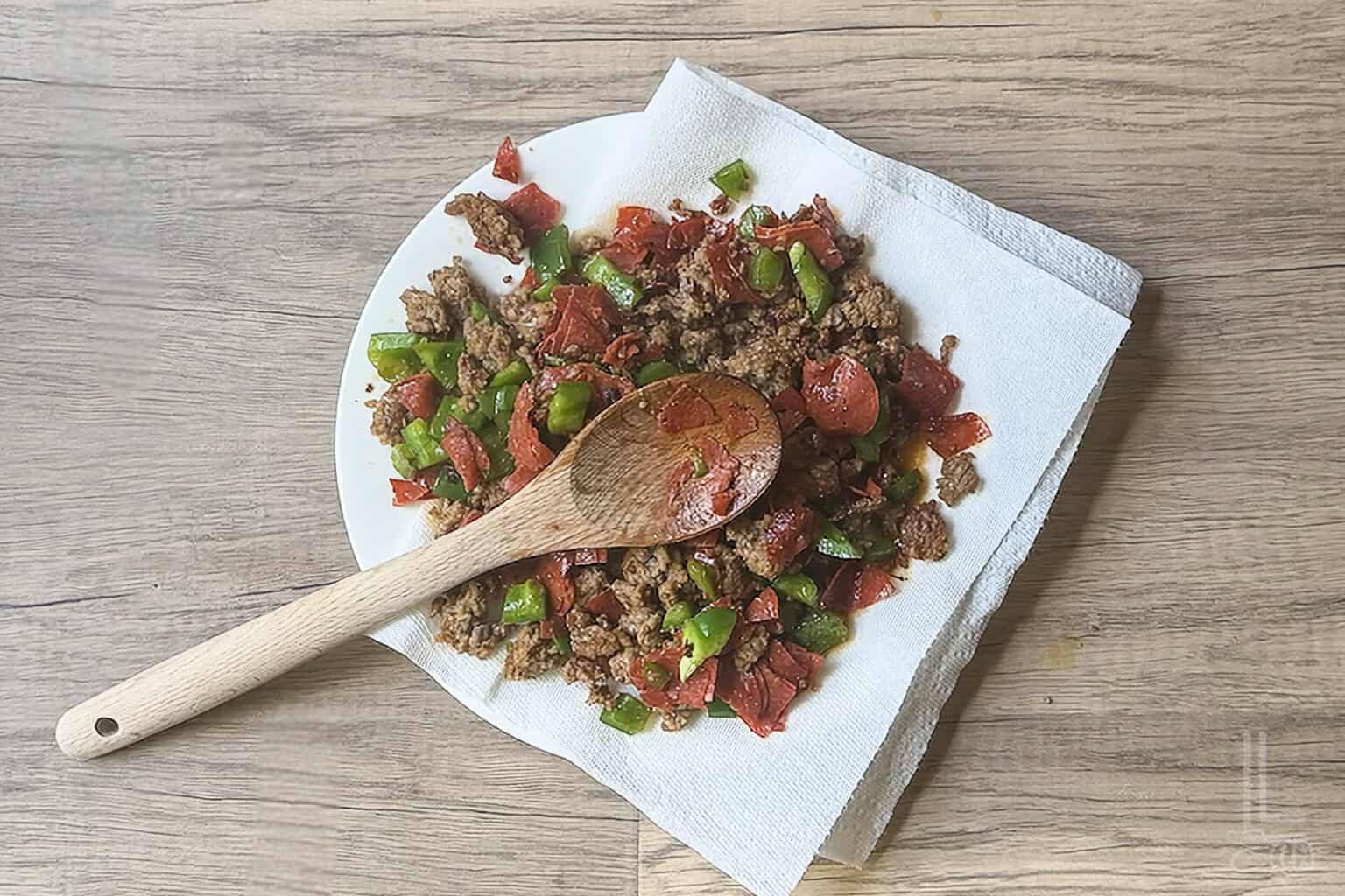 Cooked Italian sausage, pepperoni, and green bell peppers on a plate with a wooden spoon on a paper towel.