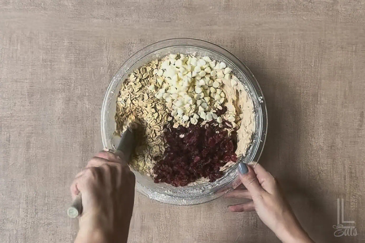 Mixing oatmeal, cranberries, and white chocolate into cookie dough in a glass bowl for cookies.