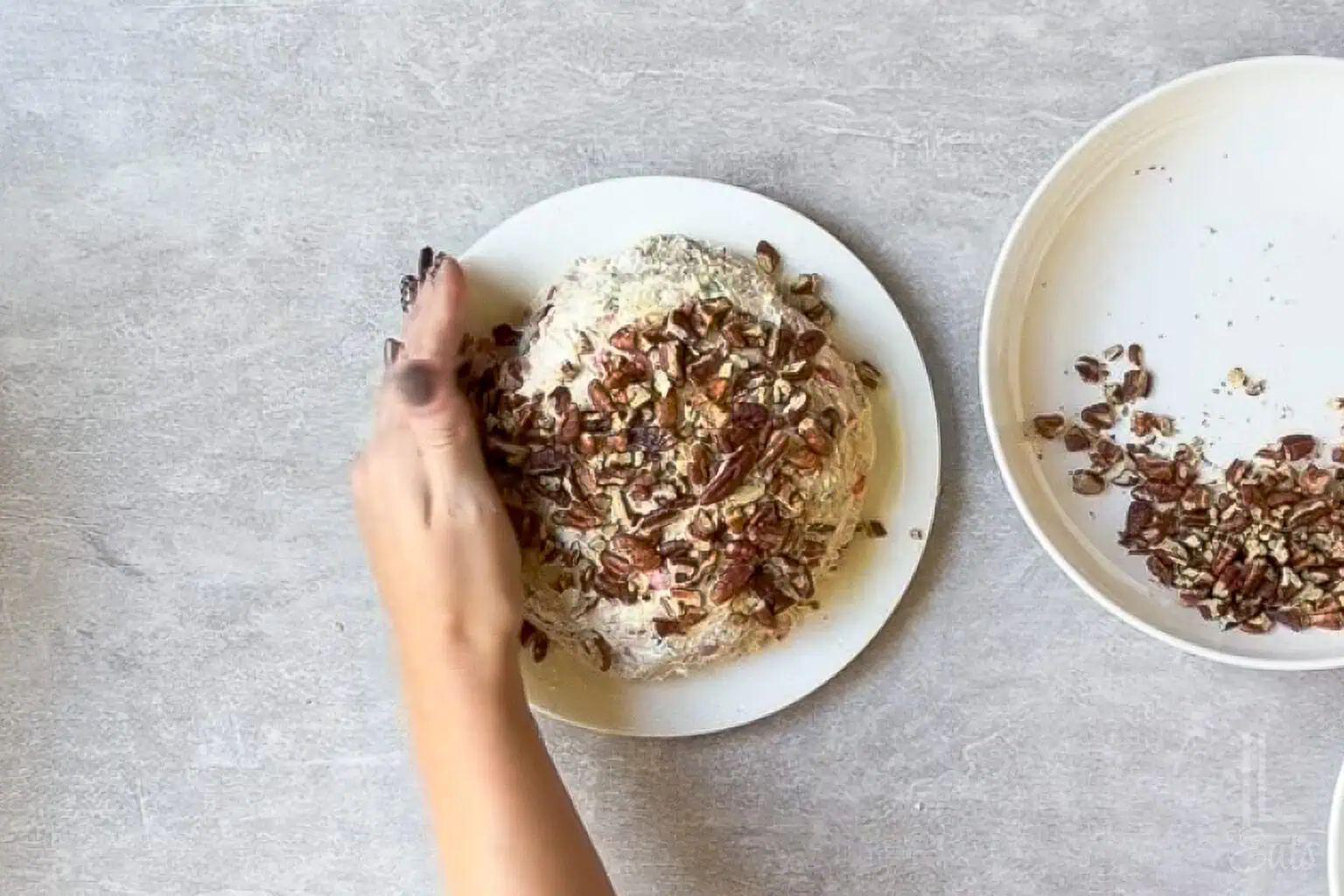 Pineapple cheese ball with bacon being prepared on a white plate, pressing pecans into the sides.