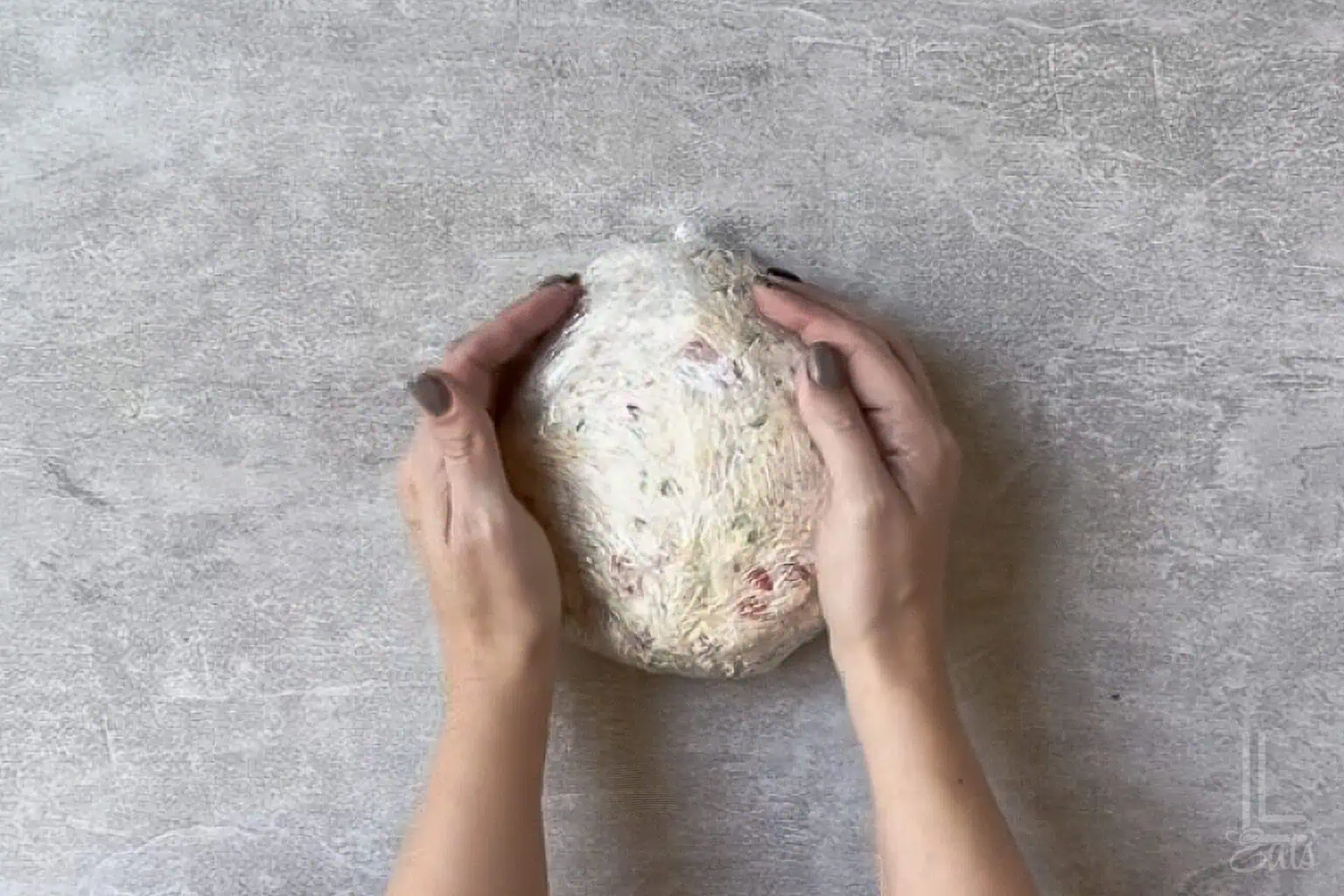 Hands shaping a pineapple cheese ball with bacon wrapped in plastic wrap.