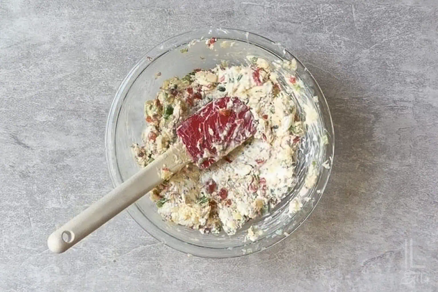 Mixing crushed pineapple, cream cheese, peppers, and bacon for a cheese ball in a glass bowl.